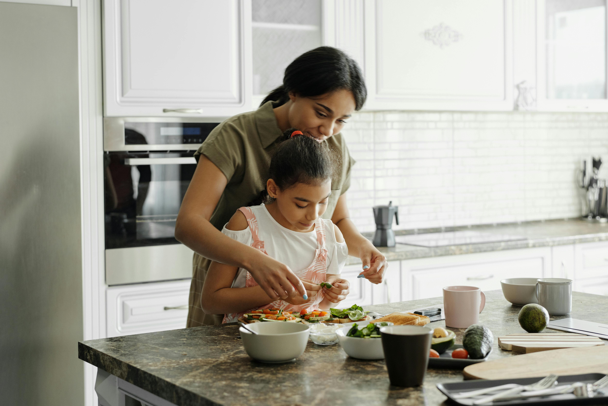 Adult and child preparing vegetables together in a bright home kitchen, with bowls, avocado and utensils on counter area