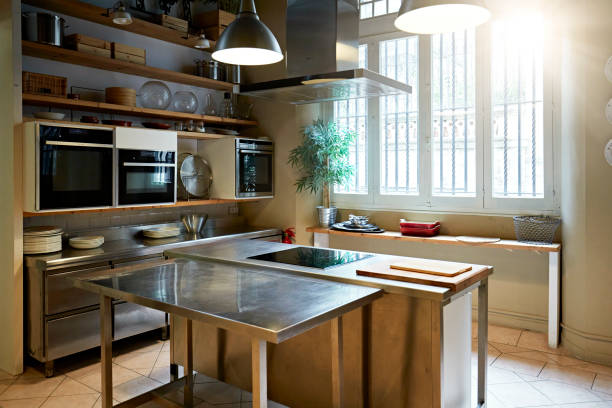 Modern kitchen with stainless steel island bench, built-in ovens, wooden shelves, hanging lights, and large window letting in natural light.